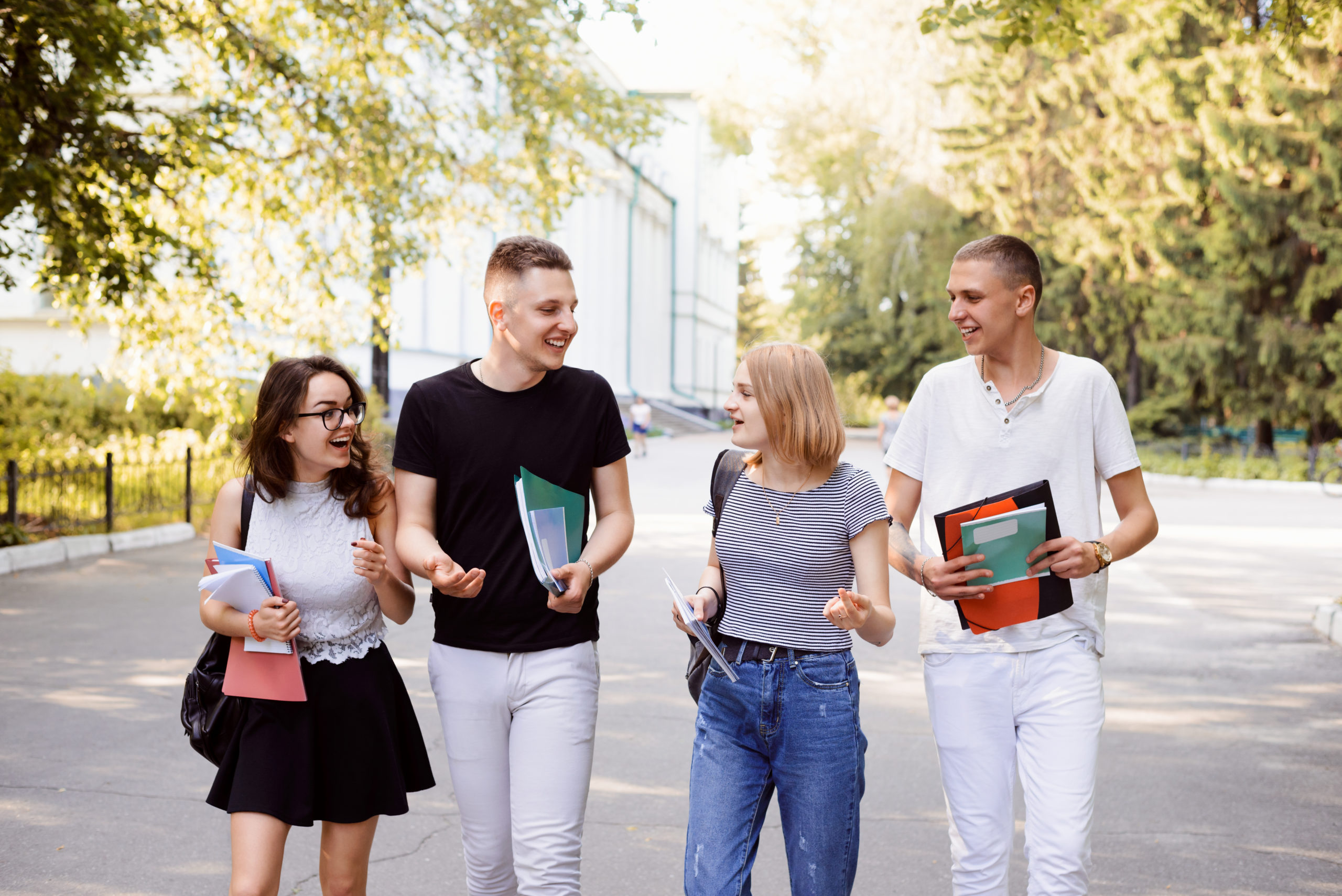 Front view of four students walking and talking in an university campus, having great free time after classes with friends, having conversation together